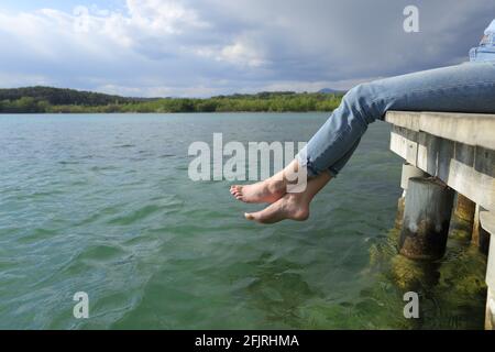 Vista laterale ravvicinata delle gambe e dei piedi di una donna riposarsi in un molo del lago Foto Stock