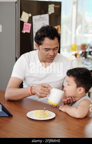 Padre che dà a suo figlio piccolo una tazza d'acqua quando stanno mangiando la colazione e guardando film animati su tablet computer Foto Stock