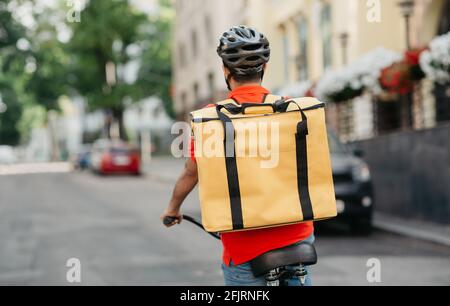 Corriere giovane maschio in t-shirt arancione, casco di sicurezza con zaino termico giallo, passeggiate in bicicletta su strada Foto Stock