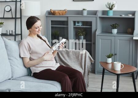Giovane donna incinta seduta sul divano guardando l'immagine radiografica e sorridendo nella stanza di casa Foto Stock