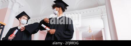 Vista ad angolo basso di studenti interracial in maschere mediche che tengono diplomi e facendo gomito bump, banner Foto Stock