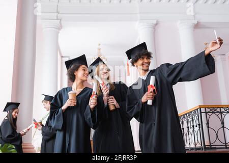 Allegri laureati multietnici con diplomi e tazze di carta che prendono selfie Foto Stock