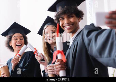 Allegri laureati interrazziali con tazze di carta e diplomi che prendono selfie sullo smartphone Foto Stock