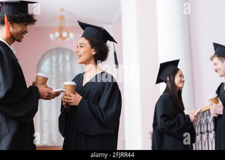 Sorridente diplomati afroamericani con smartphone e tazze di carta che parlano all'università Foto Stock