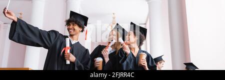 Sorridente laureati multietnici con tazze di carta che prendono selfie, banner Foto Stock