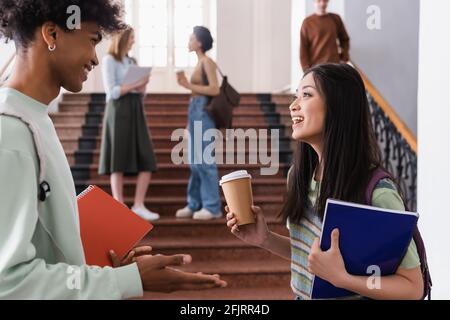 Studente asiatico con bevanda da asporto che parla con un amico afroamericano con notebook Foto Stock