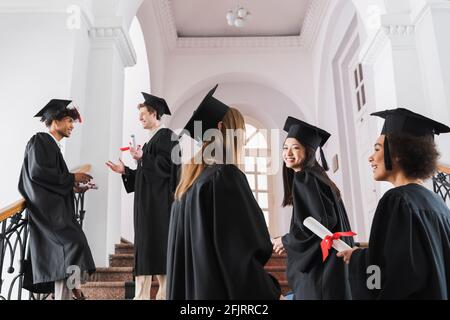 Laureati interrazziali in abiti accademici e cappelli che parlano all'università Foto Stock