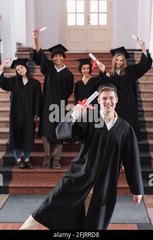 Felice laureato tenuto diploma vicino amici interrazziali in abiti accademici Foto Stock