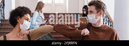 Studenti interrazziali in maschere mediche facendo gomito bump, banner Foto Stock