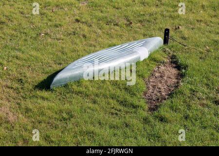 Vecchia barca in plastica dilapidata grigio chiaro con riparata sezione capovolta e lasciata legata a un piccolo metallo arrugginito palo sul lato della riva del fiume Foto Stock