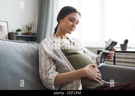 Donna incinta seduta sul divano e guardando l'immagine ecografica del suo bambino in camera Foto Stock
