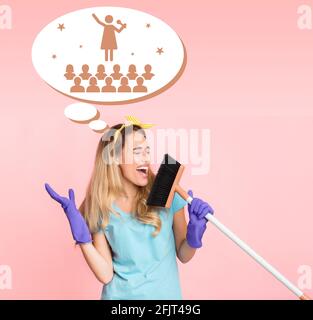 Giovane casalinga bionda che canta, usando scopa come microfono, sognando di esibirsi sul palco, sfondo rosa dello studio Foto Stock