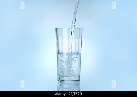 il vetro trasparente è riempito di acqua da un flusso di liquido, drinkware su uno sfondo blu con spazio di copia. Foto Stock