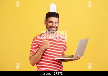 Eccitato uomo bearded con la casa di carta sulla testa che tiene il laptop che mostra i pollici in su, guardando la macchina fotografica con il sorriso toothy, servizio immobiliare, noleggio online Foto Stock