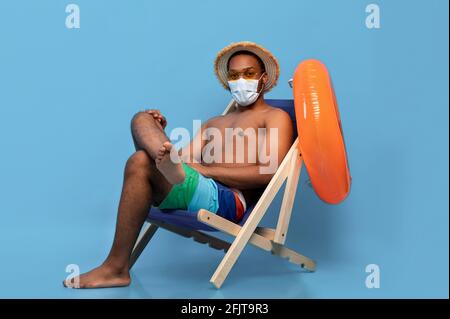 Restrizioni di viaggio durante l'epidemia di covidio. Lunghezza completa di ragazzo nero in maschera seduta in poltrona, sfondo blu Foto Stock