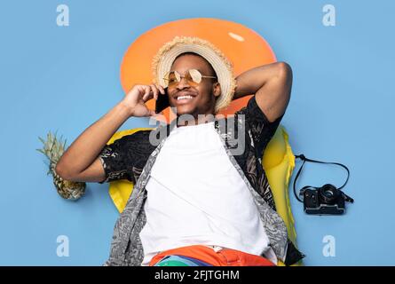 Funky ragazzo nero in abiti estivi alla moda sdraiato su gonfiabile lilo, parlando su smartphone su sfondo blu, vista dall'alto Foto Stock