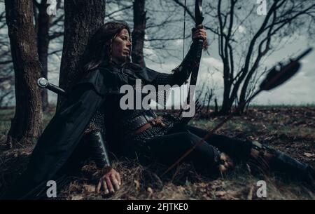 Donna in catena posta in immagine di guerriero medievale si siede e riposa vicino albero tra la foresta. Foto Stock