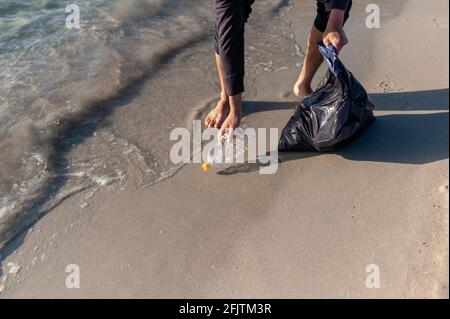 Uomo irriconoscibile che pulisce la spiaggia dai rifiuti. Primo piano su piedi e mano raccogliendo una bottiglia di plastica vuota dal mare e mettendo sulla ga Foto Stock