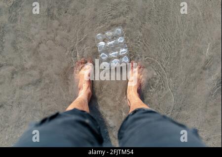 Vista dall'alto i piedi dell'uomo sulla spiaggia circondata da rifiuti di plastica. Foto Stock