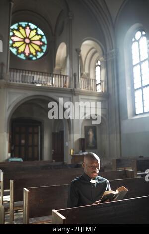 Sacerdote africano seduto sulla panchina in chiesa che legge il Bibbia e preghiera Foto Stock