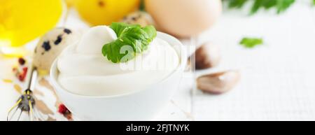 Salsa tradizionale maionese in ciotola bianca in ceramica e ingredienti per la sua preparazione su sfondo bianco di legno. Messa a fuoco selettiva. Foto Stock