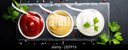 Set di tre salse - maionese, senape e ketchup in ciotole bianche in ceramica su fondo di pietra nera o calcestruzzo. Messa a fuoco selettiva. Vista dall'alto. Foto Stock