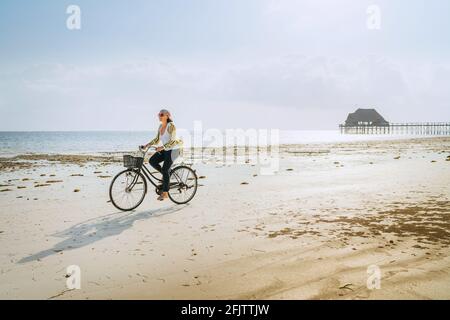 Giovane donna vestita abiti estivi leggeri a cavallo vecchia bicicletta d'epoca con cesto anteriore sulla sola bassa marea oceano bianco Costa di sabbia sulla spiaggia di Kiwengwa Foto Stock
