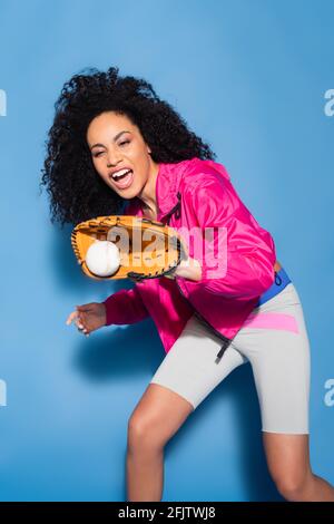 entusiasta donna afroamericana in guanto di pelle che cattura il baseball sopra blu Foto Stock