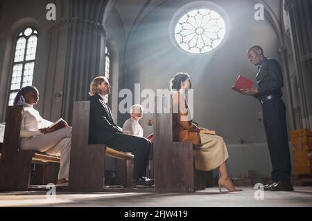 Giovane sacerdote africano che legge la Bibbia per i fedeli mentre si siedono sulla panchina della chiesa Foto Stock