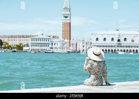 Vista da dietro elegante donna turistica solista in abito floreale con cappello seduto sul lungomare sull'isola di San Giorgio maggiore. Foto Stock