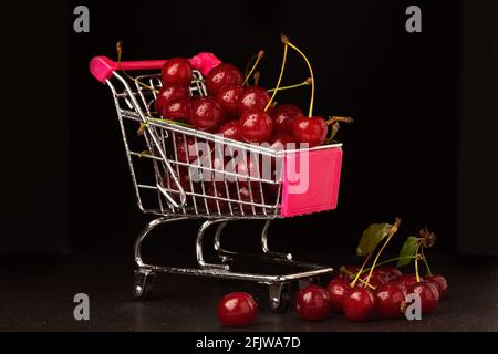 Un carrello del supermercato è riempito con ciliegie rosse mature su uno sfondo nero. Primo piano, messa a fuoco selettiva Foto Stock