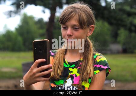 Defocus ragazza di dieci anni trova qualcosa su smartphone. Bella ragazza adolescente prende selfie mentre in piedi da sola sul parco. Blogging e live streaming Foto Stock