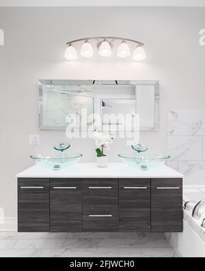 Un bel bagno vanity di legno scuro con piano di granito bianco, lavandini di vetro di cristallo, un grande specchio rettangolare, e piastrelle di marmo. Foto Stock