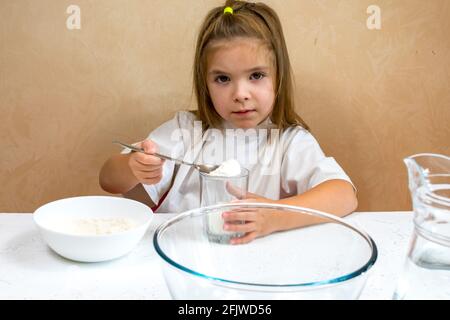 Una bambina vestita come cuoco impasta l'impasto. Cucina bambino stile di vita concetto. Il capretto ama, ha divertimento, studia e gioca nella cucina Foto Stock