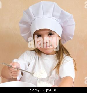 Una bambina vestita come cuoco impasta l'impasto. Cucina bambino stile di vita concetto. Il capretto ama, ha divertimento, studia e gioca nella cucina Foto Stock