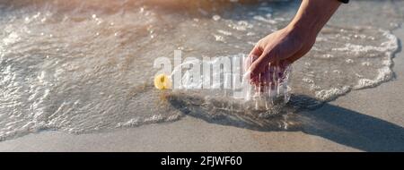 Banner orizzontale o testata uomo irriconoscibile che pulisce la spiaggia dai rifiuti. Mano ravvicinata raccolta di una bottiglia di plastica vuota dalla costa. Foto Stock