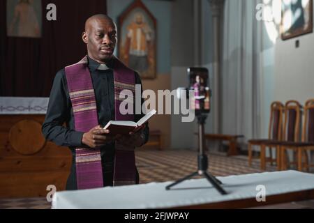 Sacerdote africano in piedi di fronte alla fotocamera sul cellulare Telefonare e leggere la Bibbia online nella chiesa Foto Stock