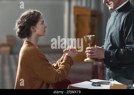 Donna fedele seduta davanti al sacerdote e pregante mentre tiene il bicchiere Foto Stock