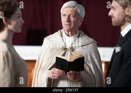Sacerdote senior che legge le preghiere e sposa la giovane coppia nella chiesa Foto Stock