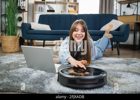 Felice giovane donna in abbigliamento casual che usa l'aspirapolvere robot per le faccende mentre lavora su un computer portatile. Donna sorridente sdraiata sul pavimento con tecnologia moderna. Foto Stock