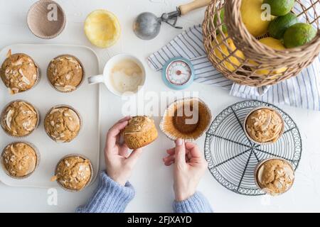 Donna che tiene una tazzina da forno in una mano e il muffin appena sfornato senza carta nell'altra mano su una tavola piena di muffin al cocco. Foto Stock