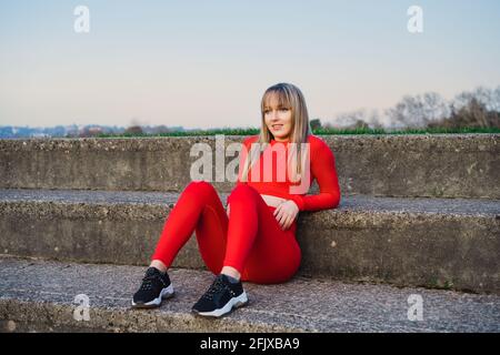 Ritratto di una donna di fitness sorridente e sicura in rosso indossano gli abiti sportivi che riposano dopo il lavoro o che corrono all'aperto al tramonto. Jogging nello stadio. Attivo Foto Stock