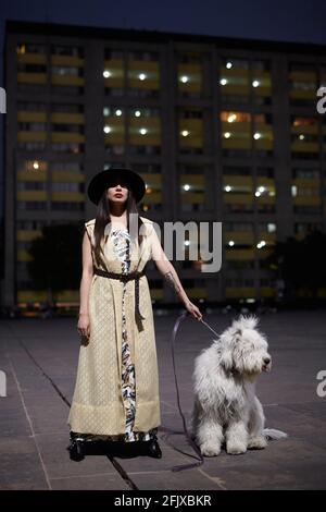elegante donna tatuata in piedi con il suo cane in città di notte Foto Stock