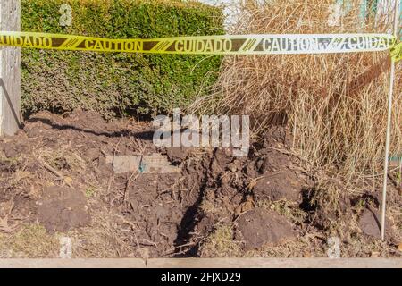 Un buco che viene scavato nel terreno per le questioni infrastrutturali tapped via da segno di attenzione in spagnolo e inglese con paesaggio in background. Foto Stock