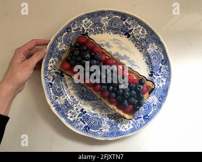 Mano della donna che tiene una crostata di mirtillo e lampone su un piatto da dessert in un banco da cucina con alcune ombre. Foto Stock