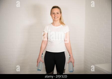 Donna bionda felice facendo esercizio di fitness con bottiglie d'acqua - allenamento sportivo di base come allenamento a casa Foto Stock