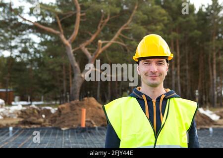 Ritratto di un costruttore maschile in un casco giallo e. giubbotto di protezione sullo sfondo della costruzione di casa-rinforzo per la fondazione Foto Stock