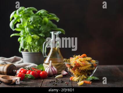 Pasta tricolore di fusilli crudi in recipiente di vetro con olio e aglio, basilico e pomodori su fondo ligneo. Spazio per il testo Foto Stock