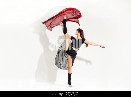 Giovane ballerina agile che esegue una posizione dinamica di pattinaggio su ghiaccio alzando la gamba in aria come lei galleggia con grazia tessuto rosso sopra la sua testa isola Foto Stock