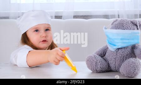 la bambina gioca un medico, fa un'iniezione. Felice bambino medico piccolo esamina un orsacchiotto. Concetto di trattamento del virus corona, iniezione durante Foto Stock